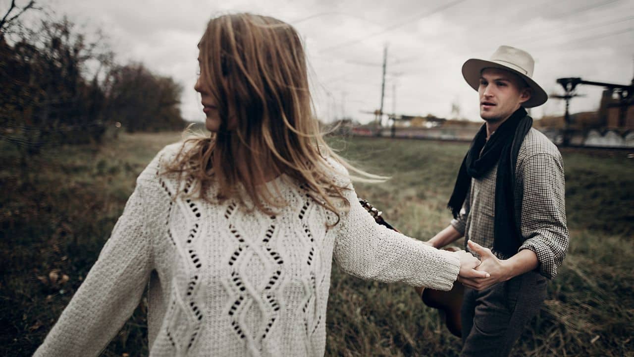 A woman in a white sweater leads a man wearing a hat through a grassy, outdoor field.