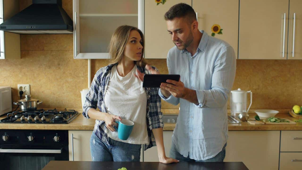 A man shows a tablet to a woman holding a blue mug in a kitchen.