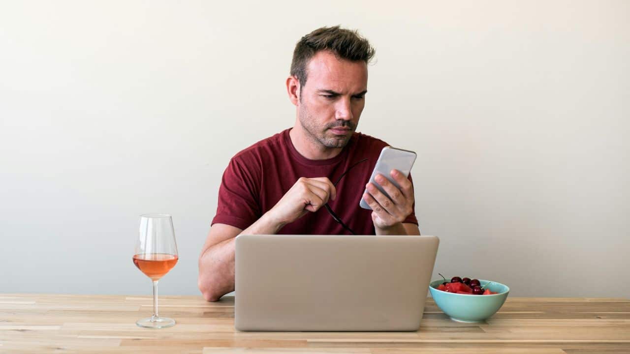 A man in a red shirt looks at a smartphone while sitting before a laptop.