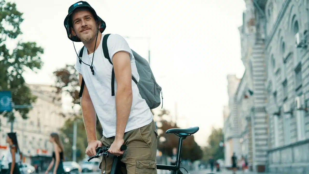 A man wearing a bucket hat and backpack stands with a bicycle on a city street.