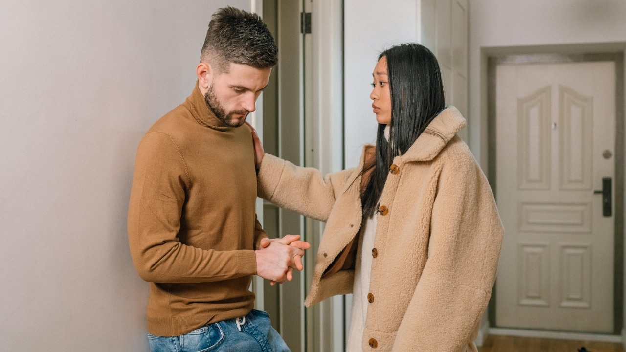 A woman in a tan coat places her hand on the shoulder of a man looking down.