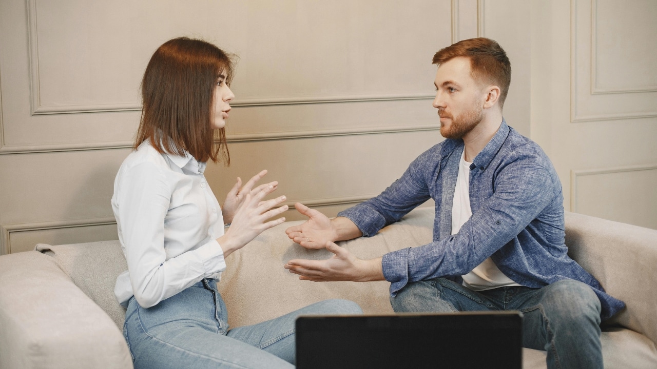 A man and woman sit on a beige sofa facing each other and gesturing with hands.