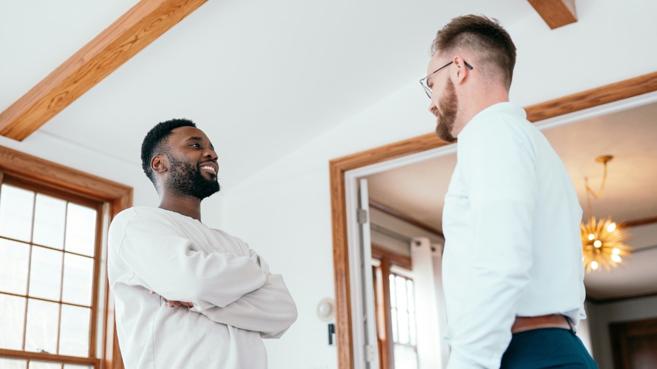 Two men stand in a room with white walls and wooden beams, looking at each other.