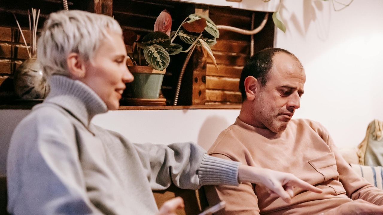 A woman in a grey turtleneck points forward while sitting next to a man.