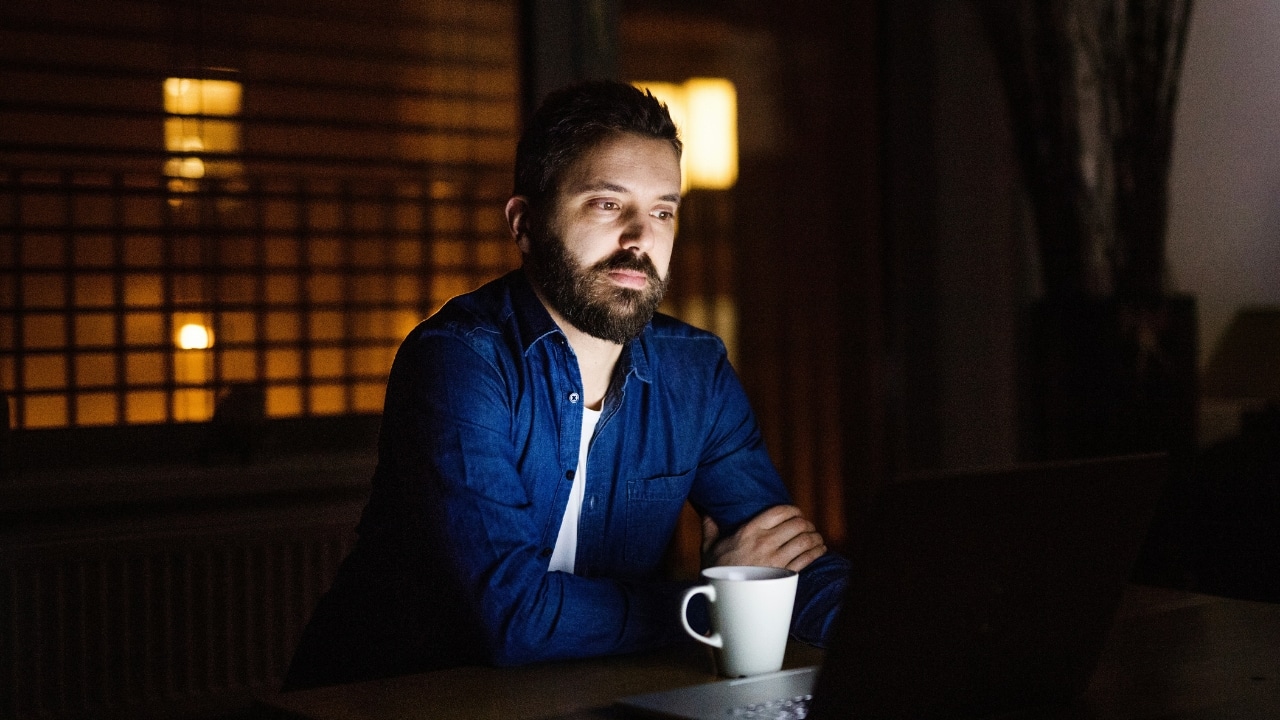 A man with a beard sits at a table with a laptop and a mug.