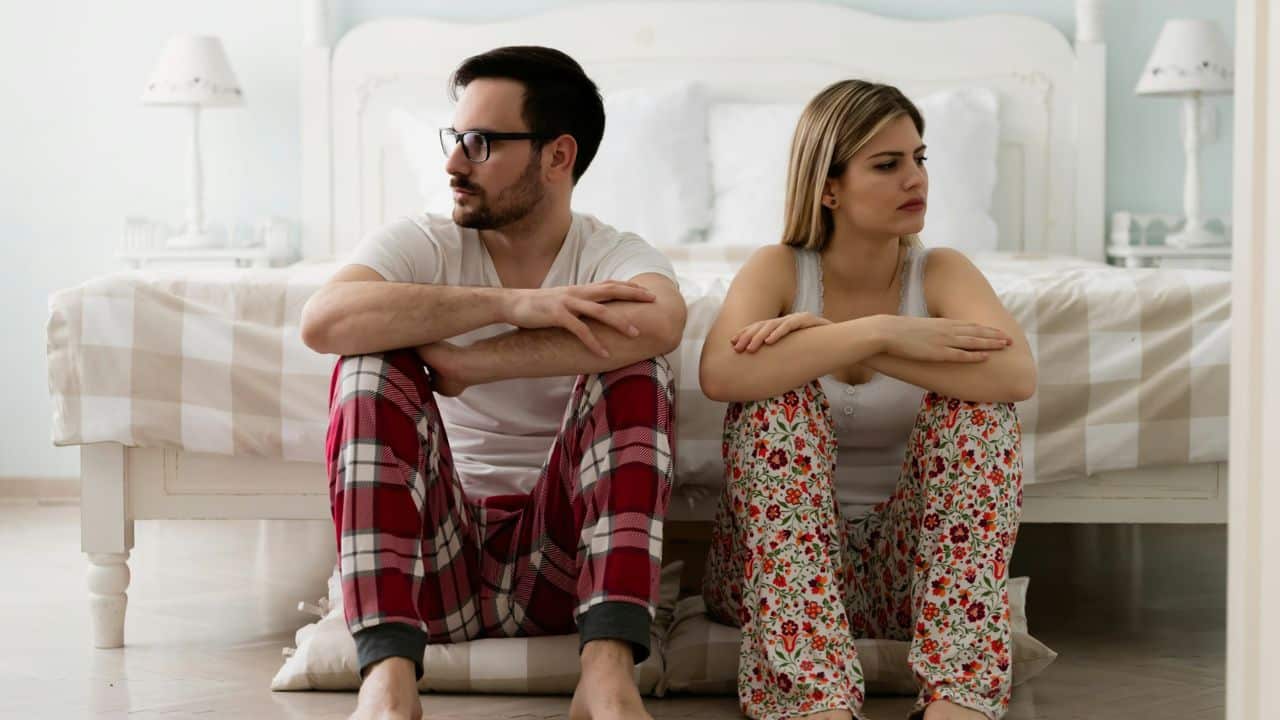 A man and woman in pajamas sit on the floor, looking away from each other.