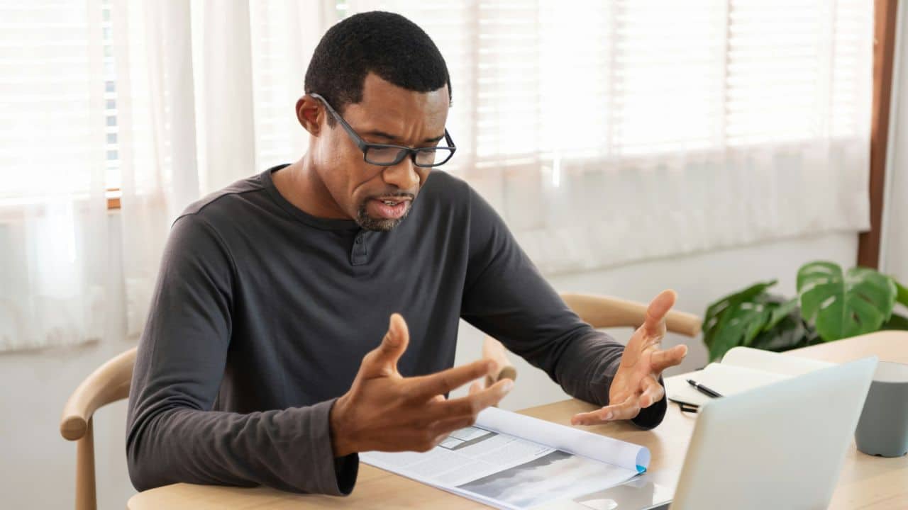 A man with glasses sits at a desk gesturing with both hands over a paper.