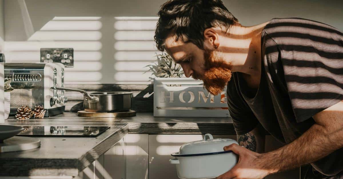 A bearded man cooking in a sunlit kitchen while holding a pot.