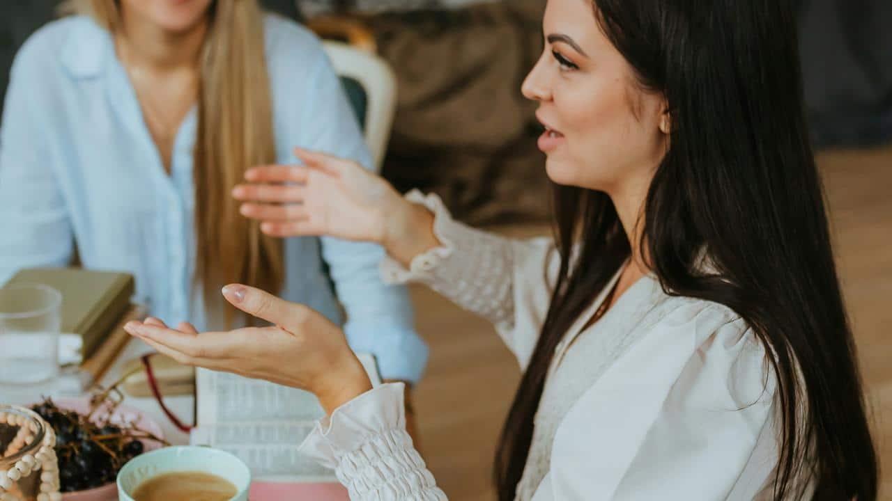 A woman gesturing while talking to another person at a table with coffee and books.