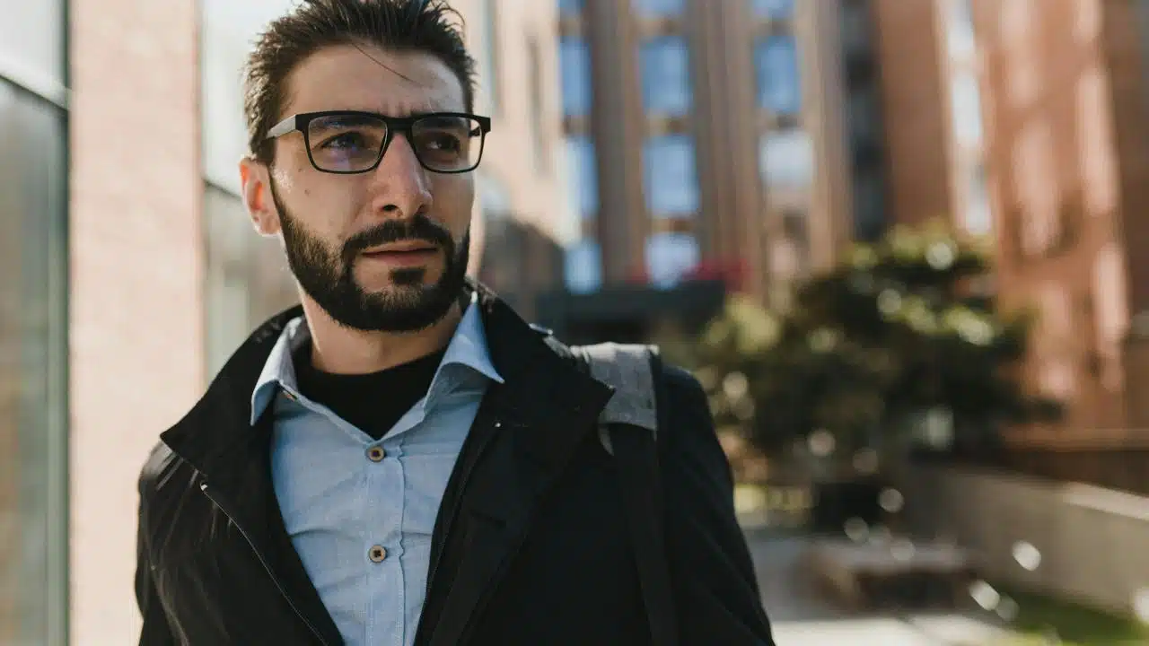 A man wearing glasses and a jacket walking outdoors in an urban area.