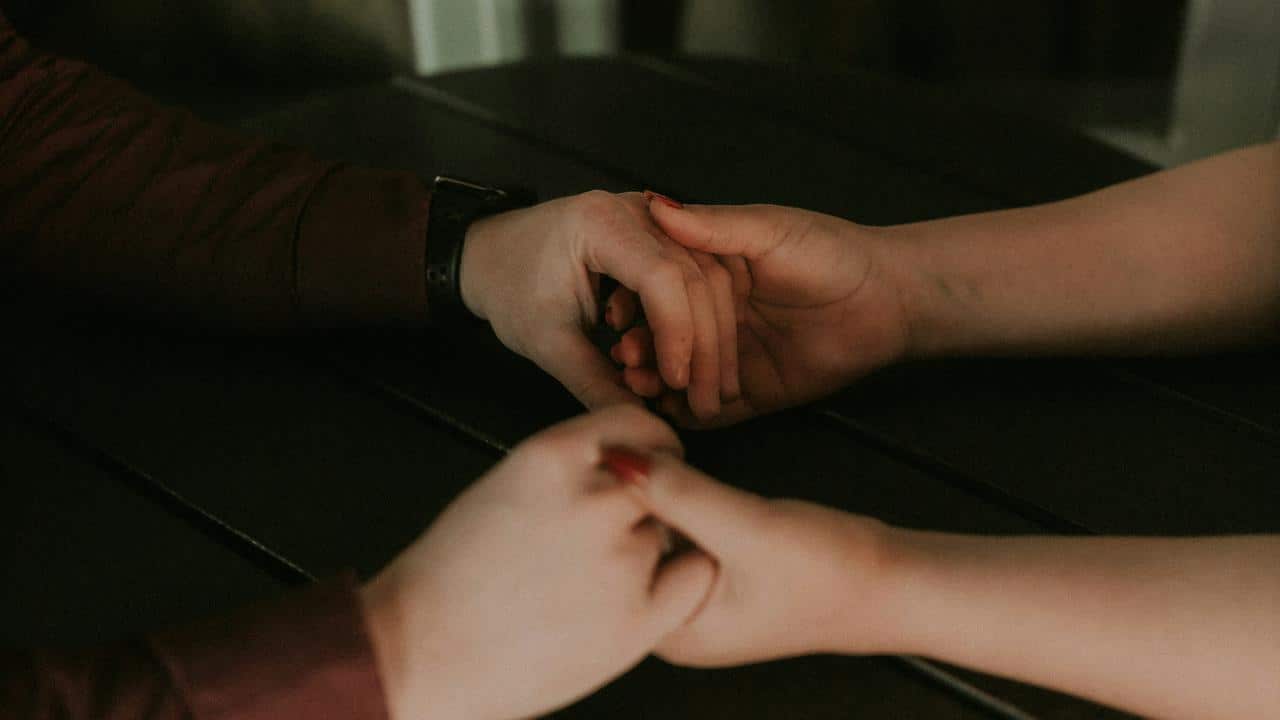 A pair of hands gently hold each other across a dark table.