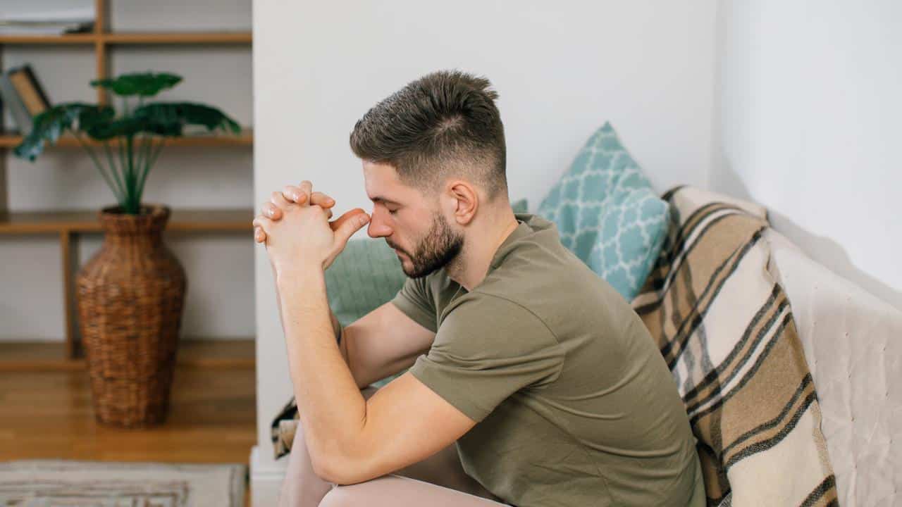 A man sitting on a couch with his hands clasped, looking thoughtful.