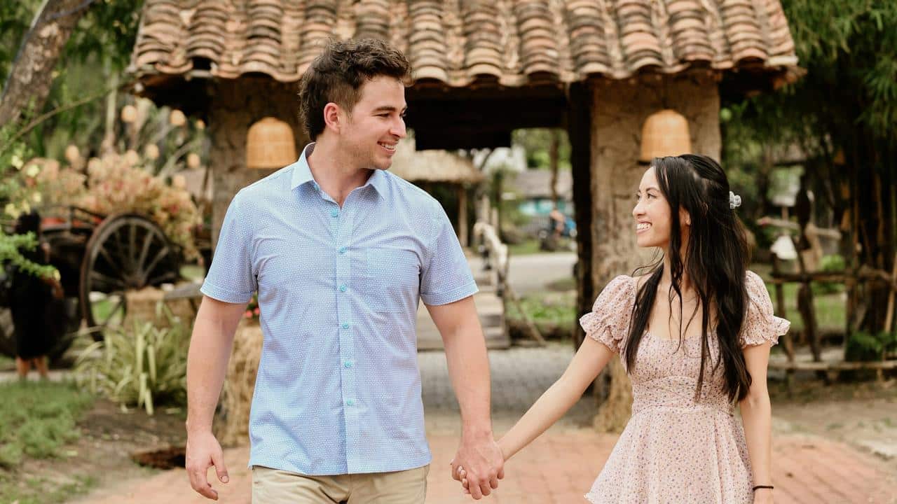 A couple holding hands and smiling while walking through a rustic outdoor setting.
