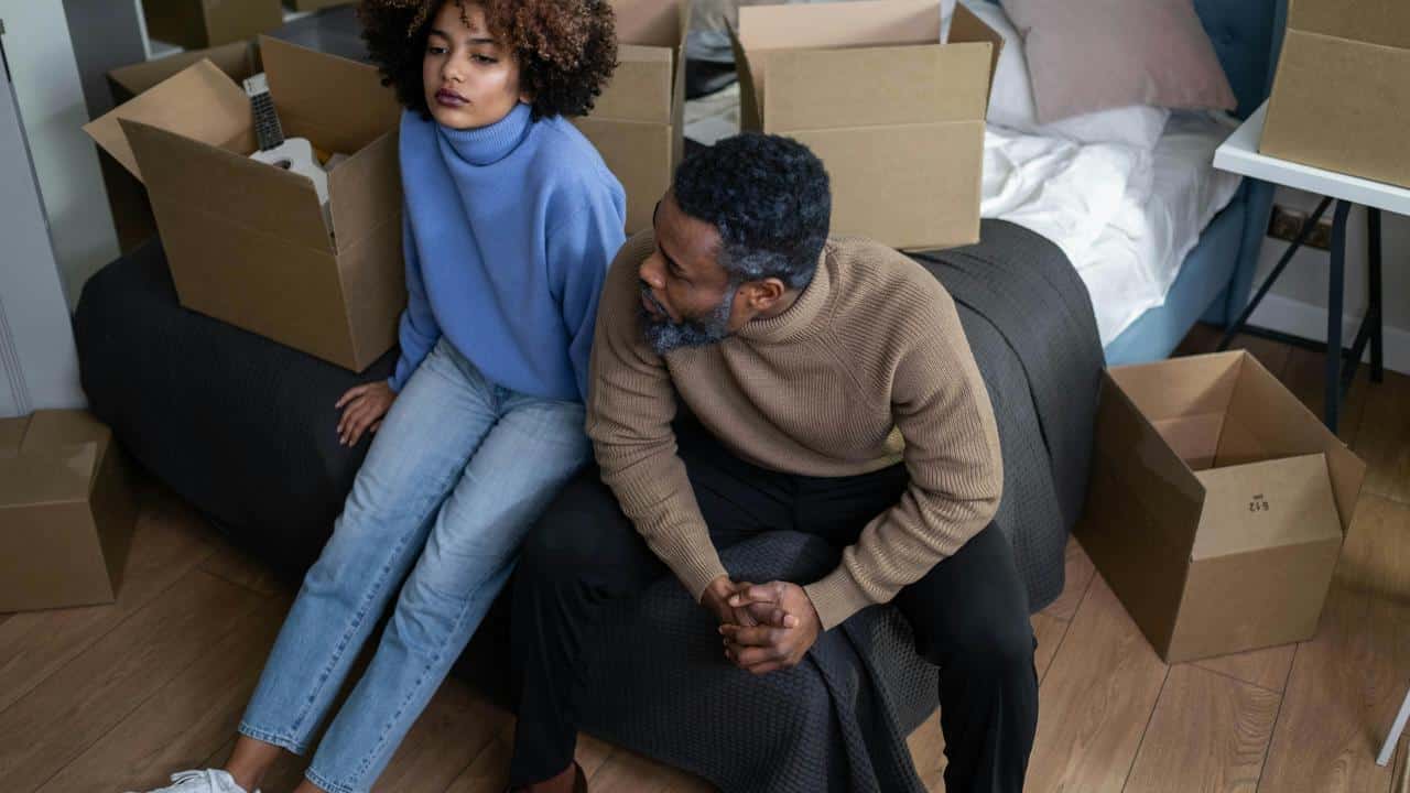 A couple sitting on a bed surrounded by moving boxes, both looking upset and distant.