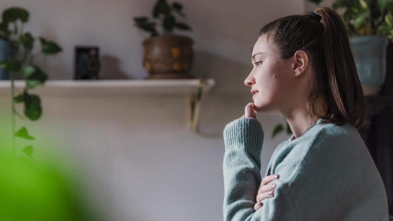 A woman sitting indoors, resting her chin on her hand while looking thoughtfully to the side.