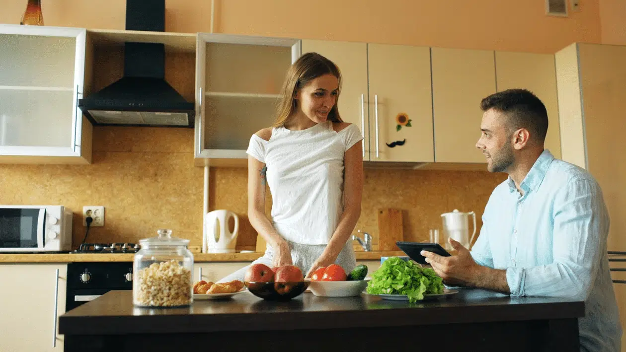 A wife cuts vegetables as the husband sits with his tablet.