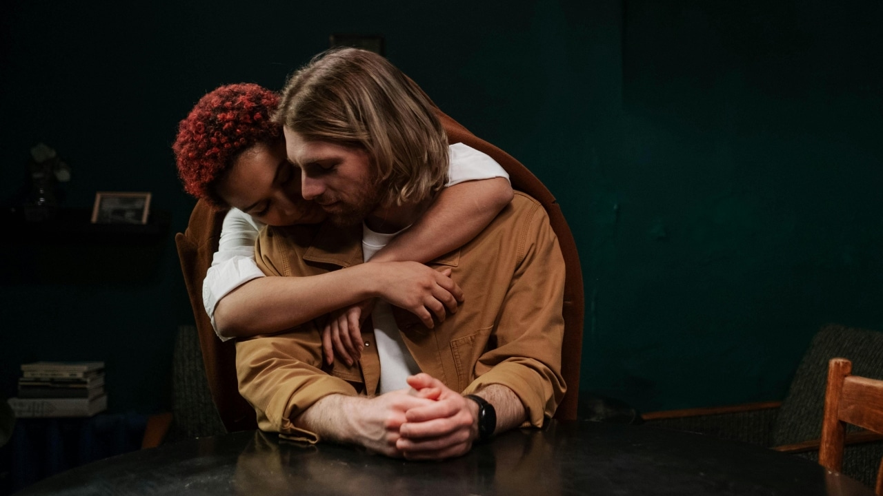 A woman with red hair hugs a man from behind while he sits at a table.