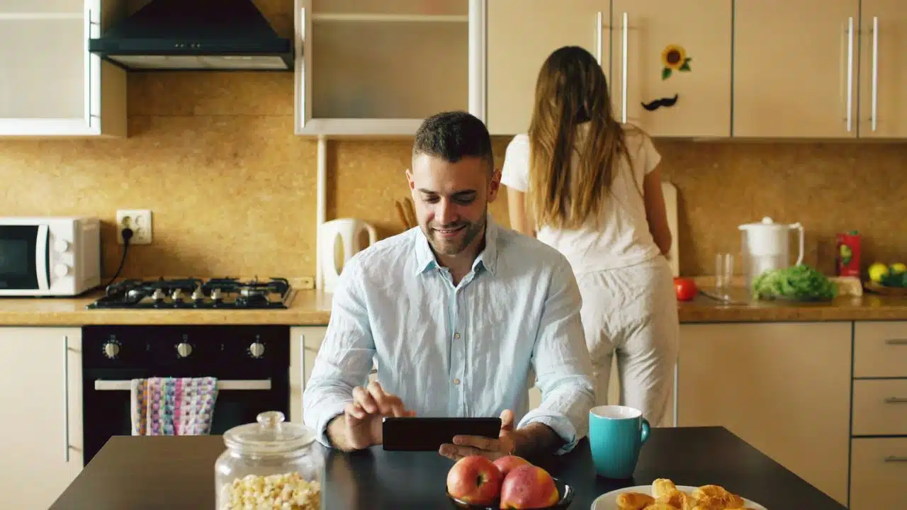 A man sits at a table using a phone while a woman works in the kitchen.