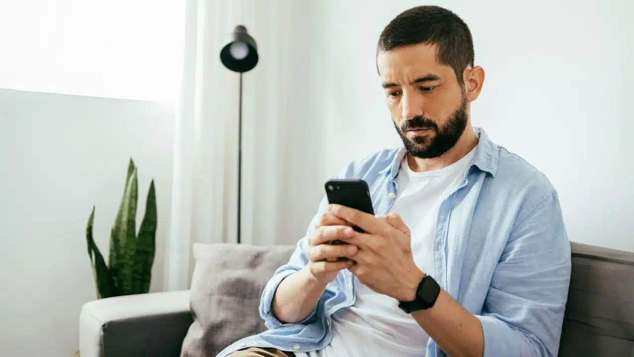 A bearded man in a light blue shirt sits on a sofa looking at his phone.