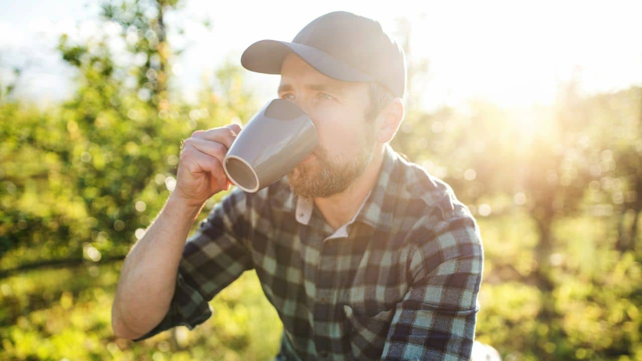 Bearded man in a cap and plaid shirt drinking from a mug outdoors in sunlight.