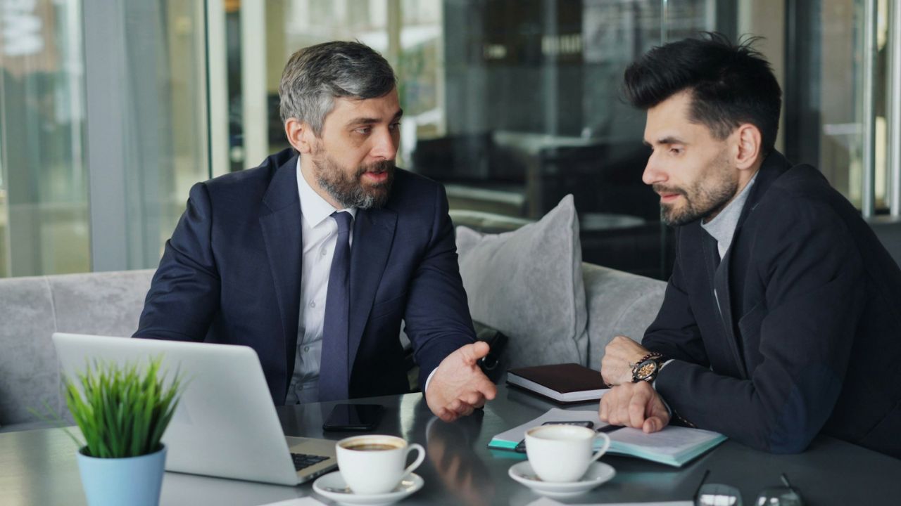 Two men in suits discussing business over coffee and a laptop in an indoor setting.
