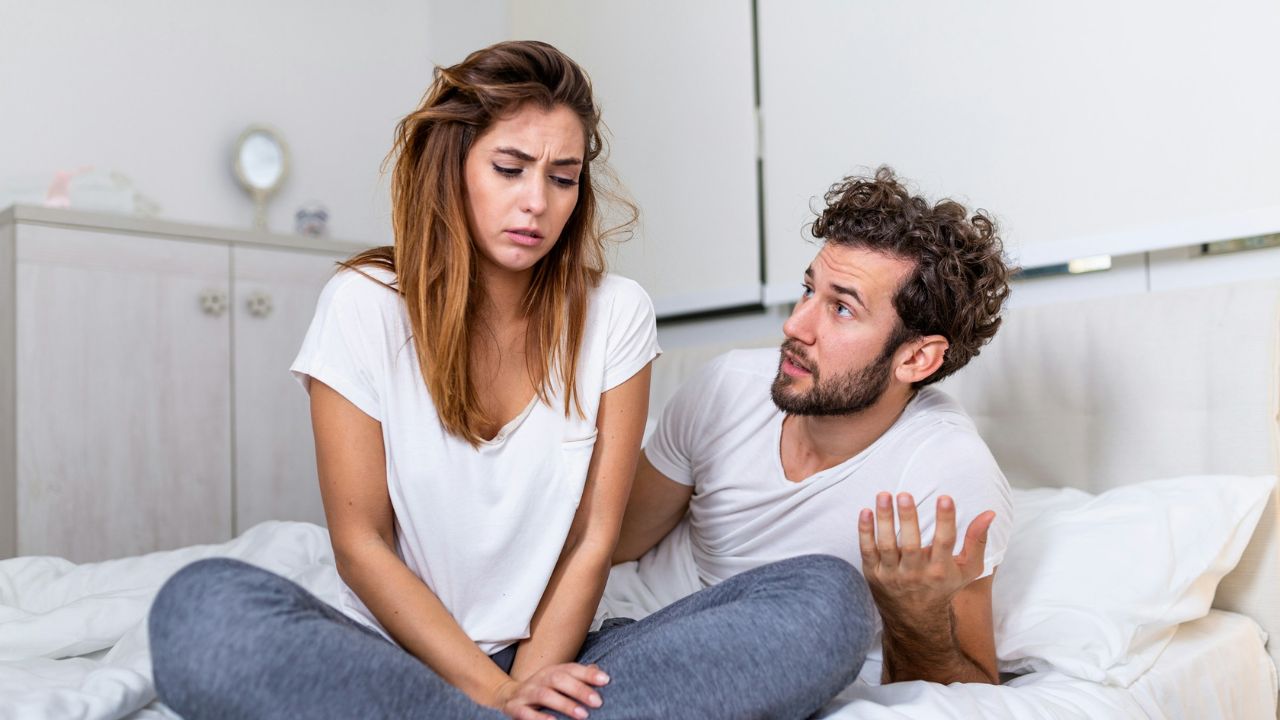 A man gesturing and talking to a sad-looking woman sitting on a bed during a disagreement.