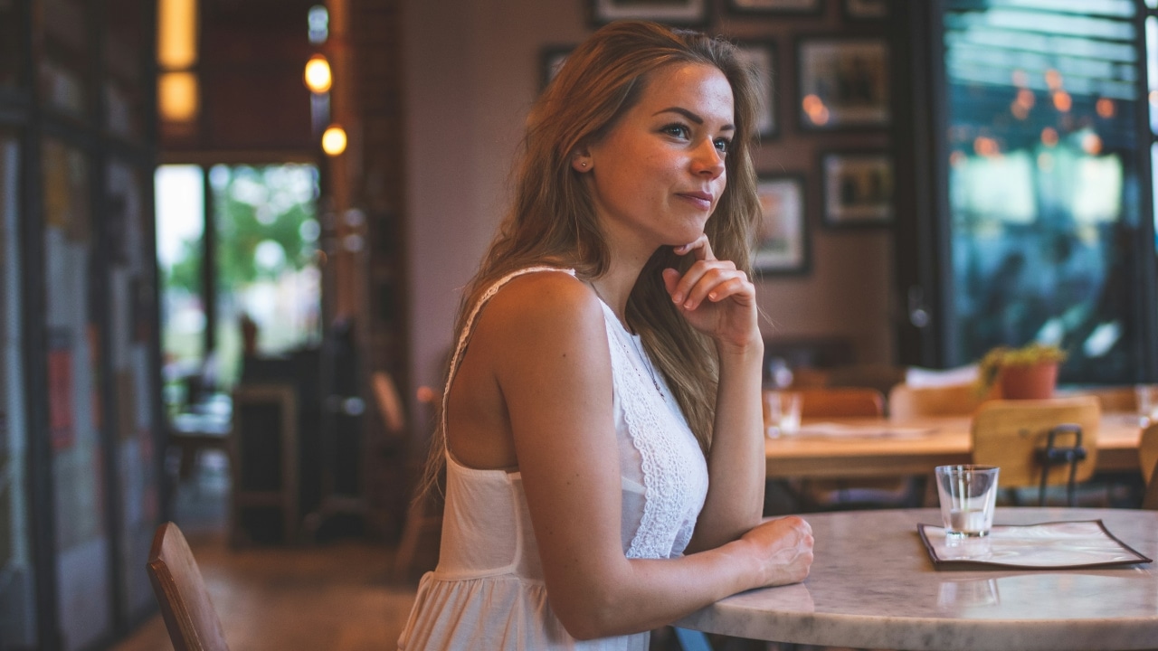 A woman in a white dress sits at a marble cafe table, looking off to the side.