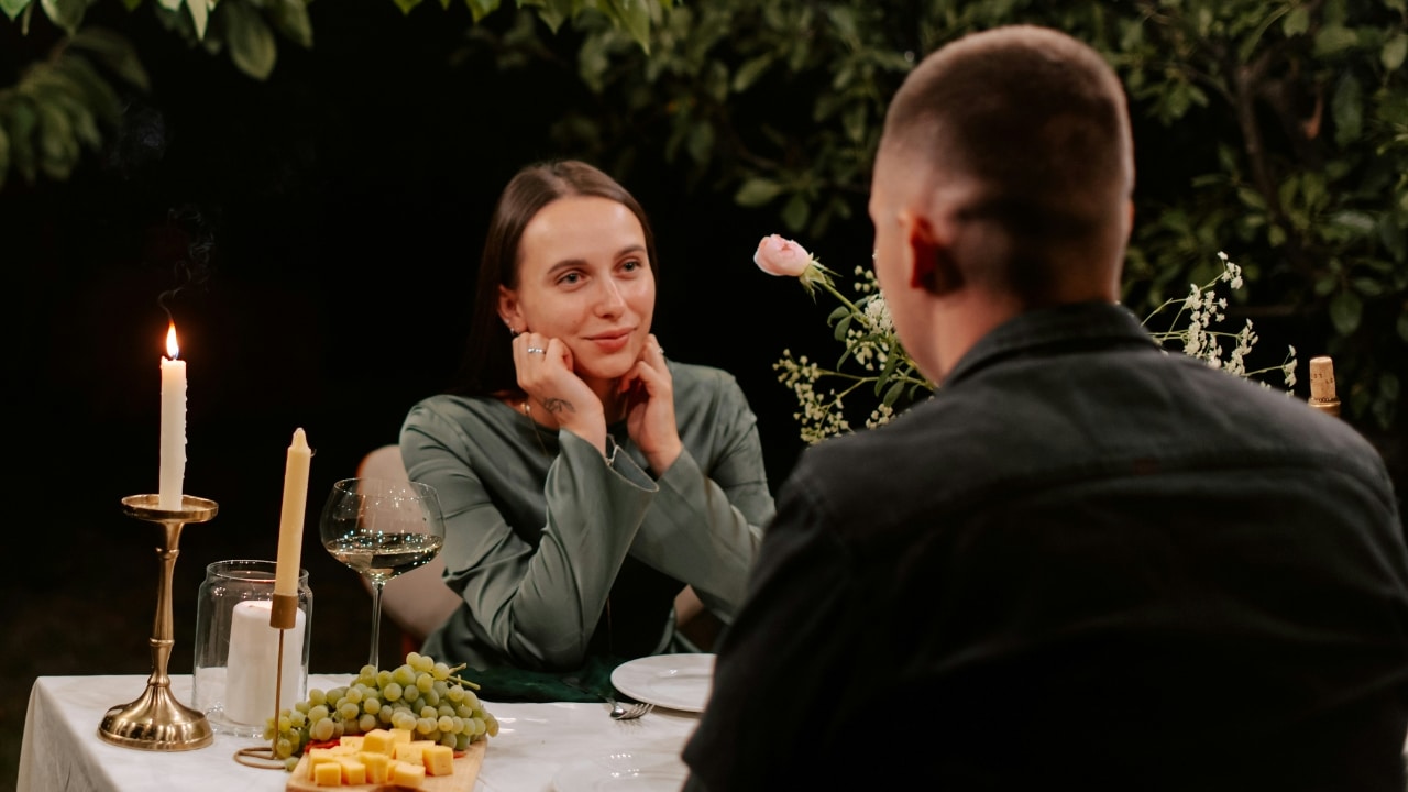 A woman rests her chin on her hands while sitting across from a man outdoors.