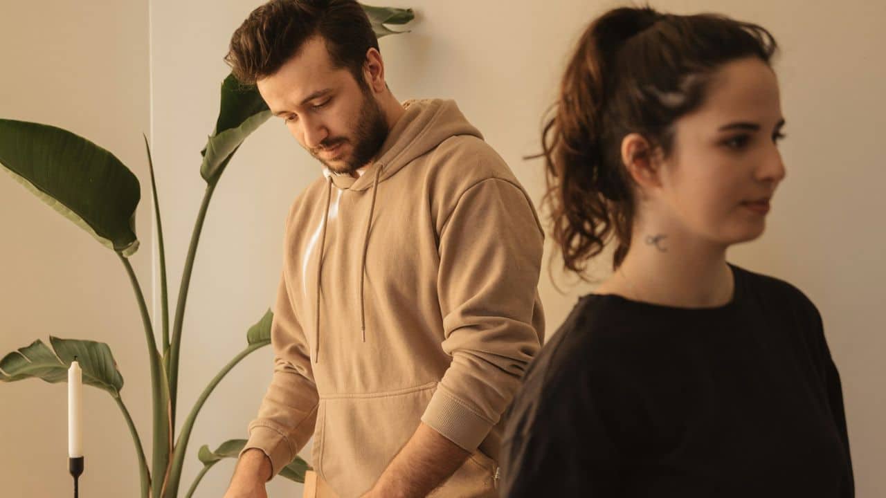 A man in a tan hoodie looks down while a woman in black stands in the foreground.
