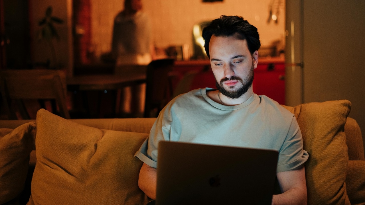A bearded man sits on a tan sofa, focused on a laptop in a warm, indoor setting.