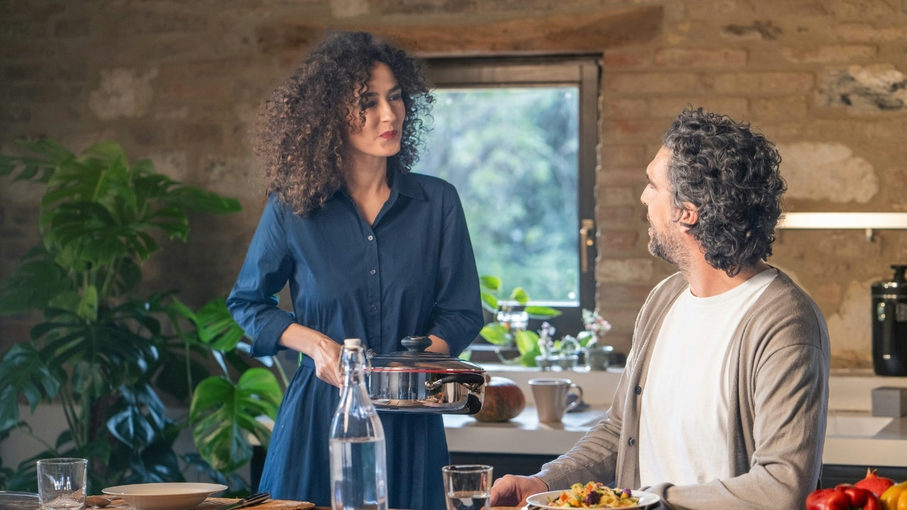 A woman holding a pot looks at a man sitting at a dining table.