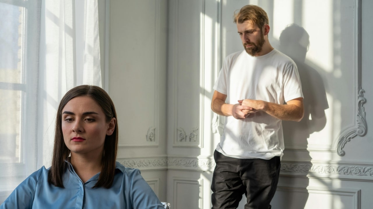 A woman sits in the foreground while a man stands against a wall behind her.