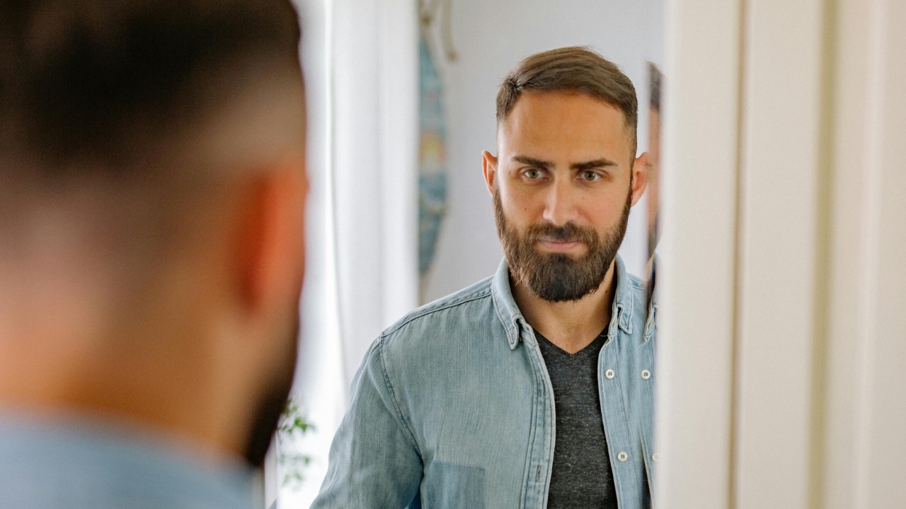 A bearded man in a denim shirt looks at his own reflection in a wall mirror.