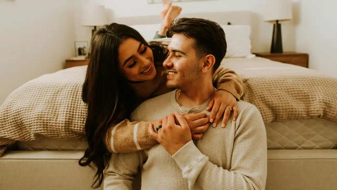 A smiling woman leans over the back of a man sitting on a bedroom floor.