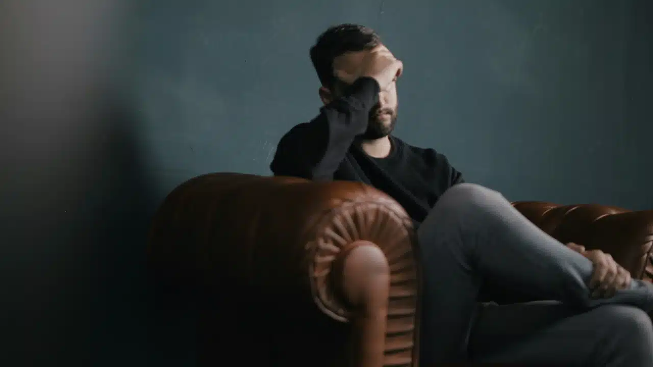 A man sits in a brown leather armchair with one hand covering his eyes and forehead.