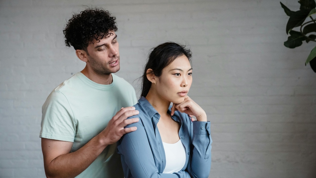 A man places his hand on the shoulder of a woman looking away with a frown.