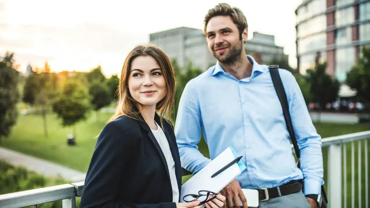 A woman in a blazer and a man in a blue shirt stand outdoors smiling.