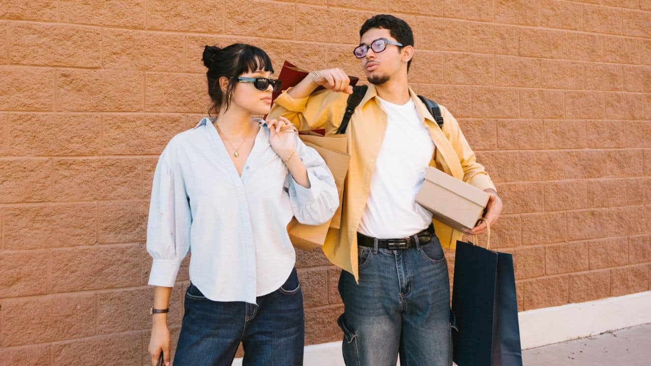 A man carrying several shopping bags and a box walks next to a woman wearing sunglasses.