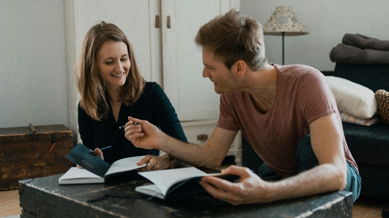 A man and woman sit on the floor by a trunk, writing in notebooks and talking.