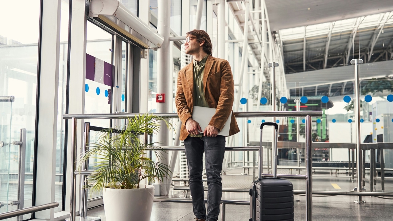 A man in a brown jacket stands in an airport holding a laptop and luggage.