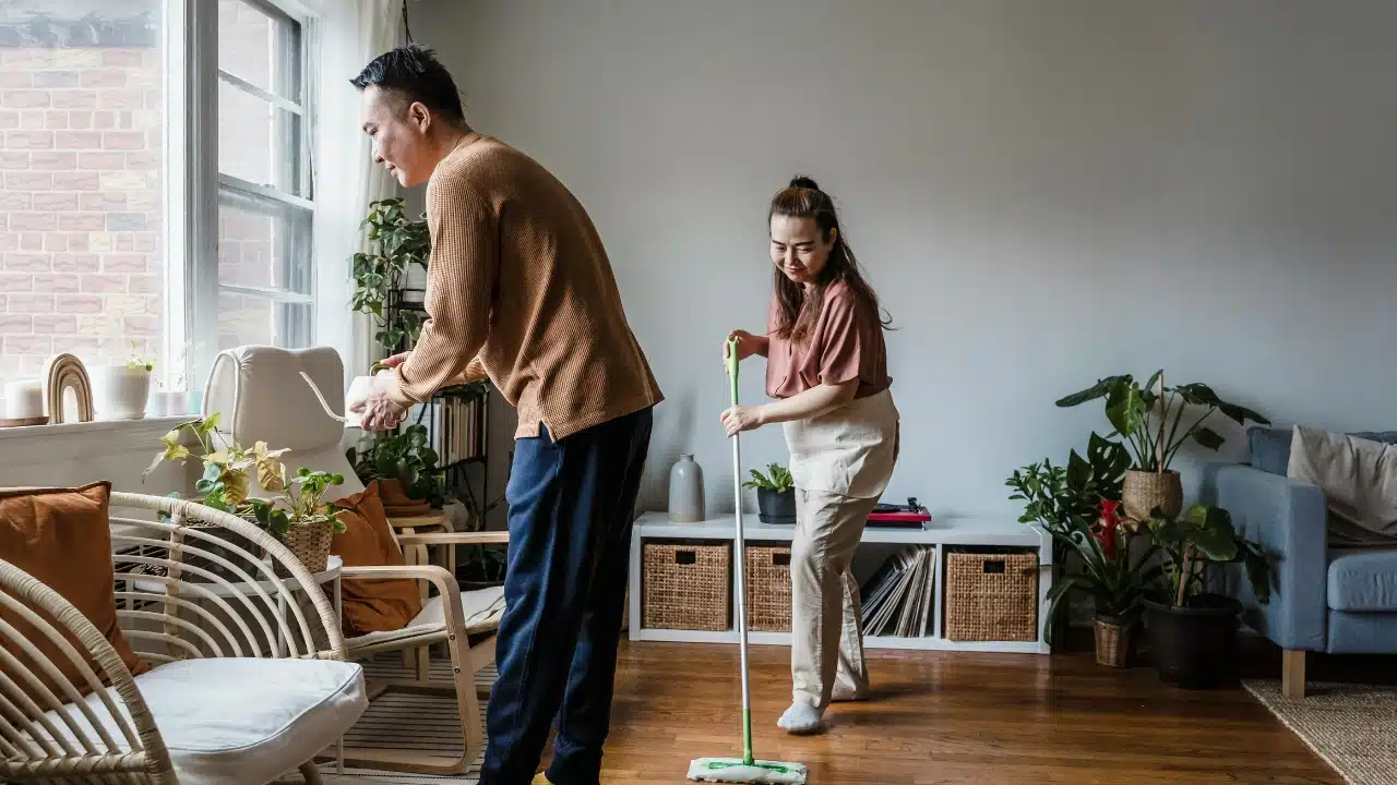 A man waters a plant by a window while a woman mops the wooden floor.