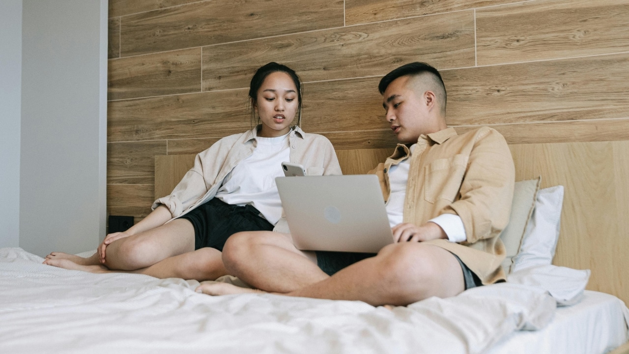A woman and a man sit on a bed while looking at a laptop together.