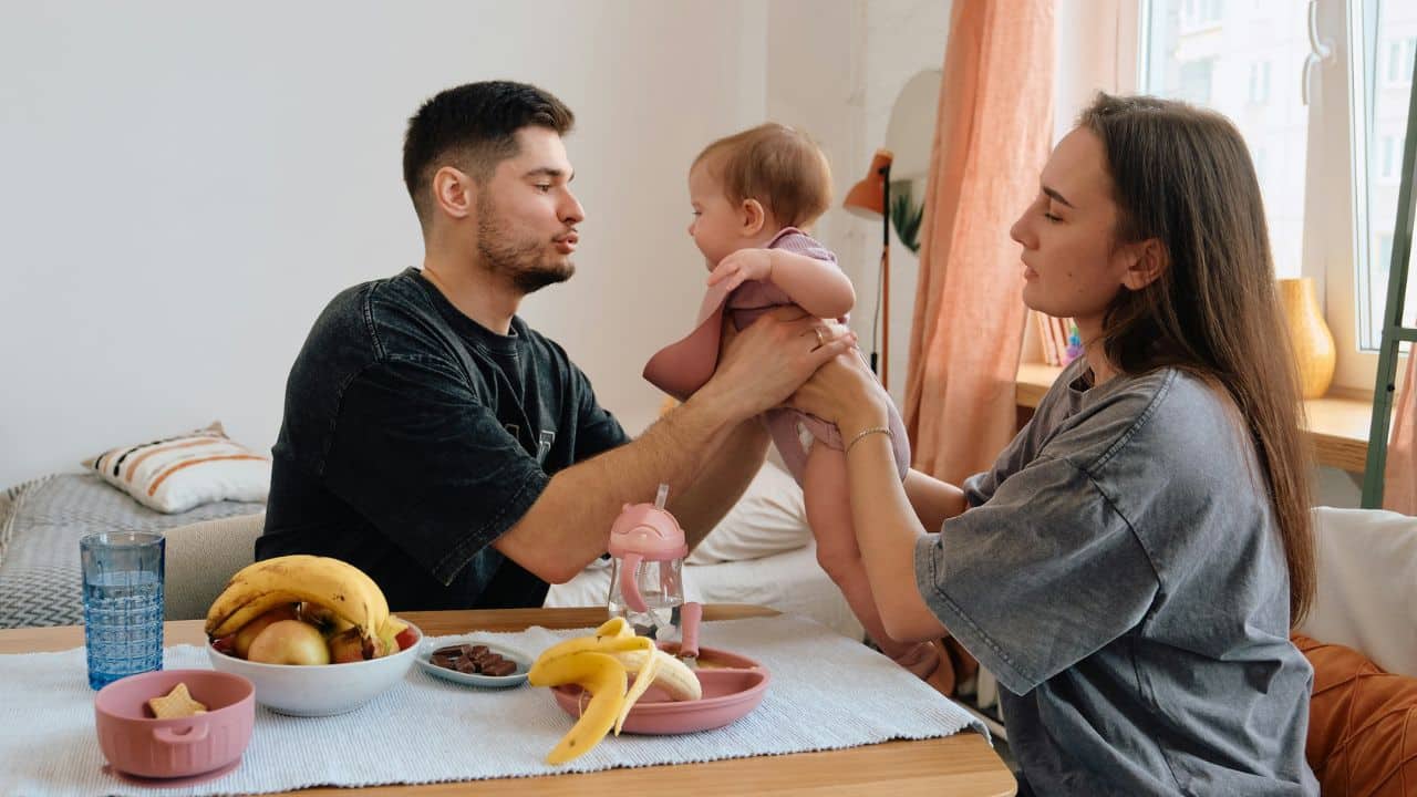 A man and woman sit at a table holding a baby during a mealtime.