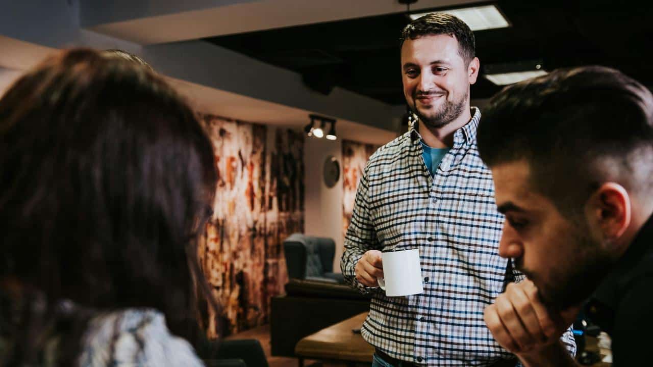 A man holding a mug and smiling while talking with two people in an office setting.