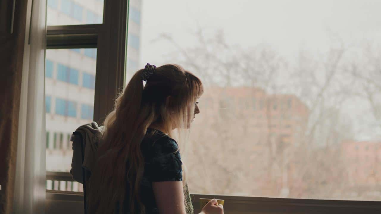 A woman standing by a window holding a cup and looking outside.