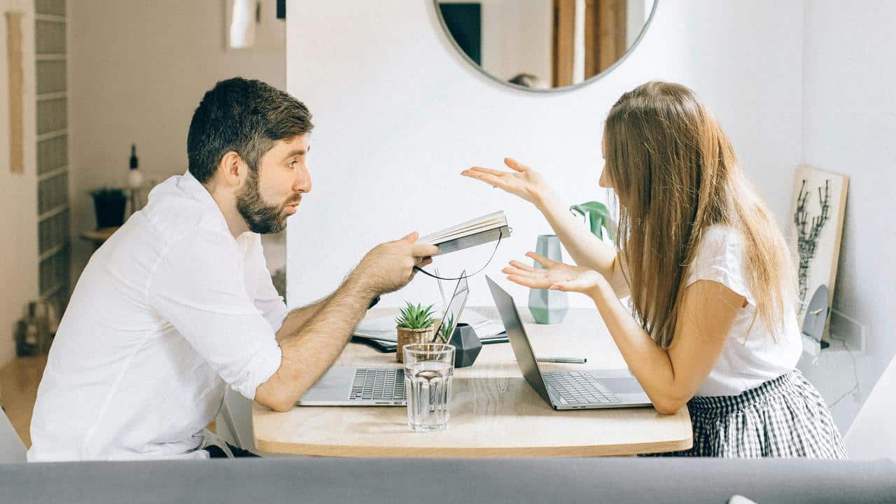 A man and woman arguing across a table with laptops in a home setting.