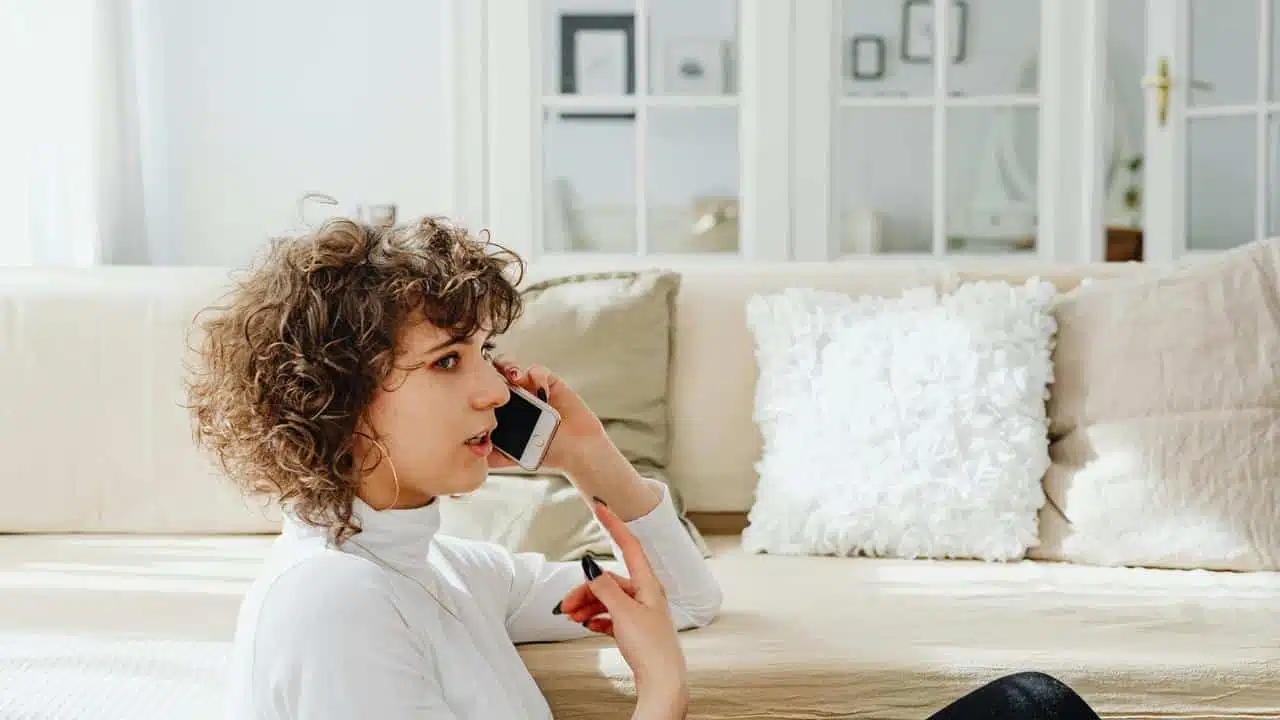 A woman sitting on the floor talking on her smartphone in a bright living room.