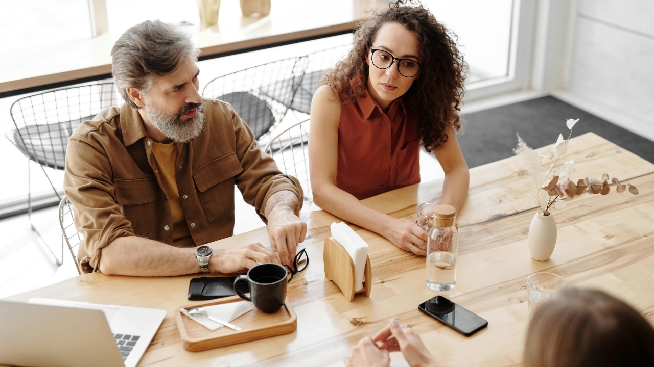A man with a beard and a woman with glasses sit at a wooden table.