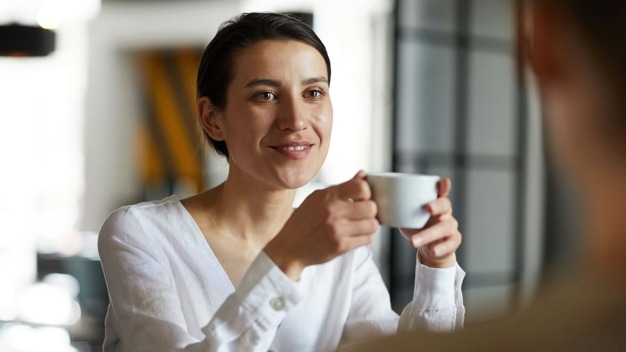 Woman in a white linen shirt holding a small white cup and smiling forward.