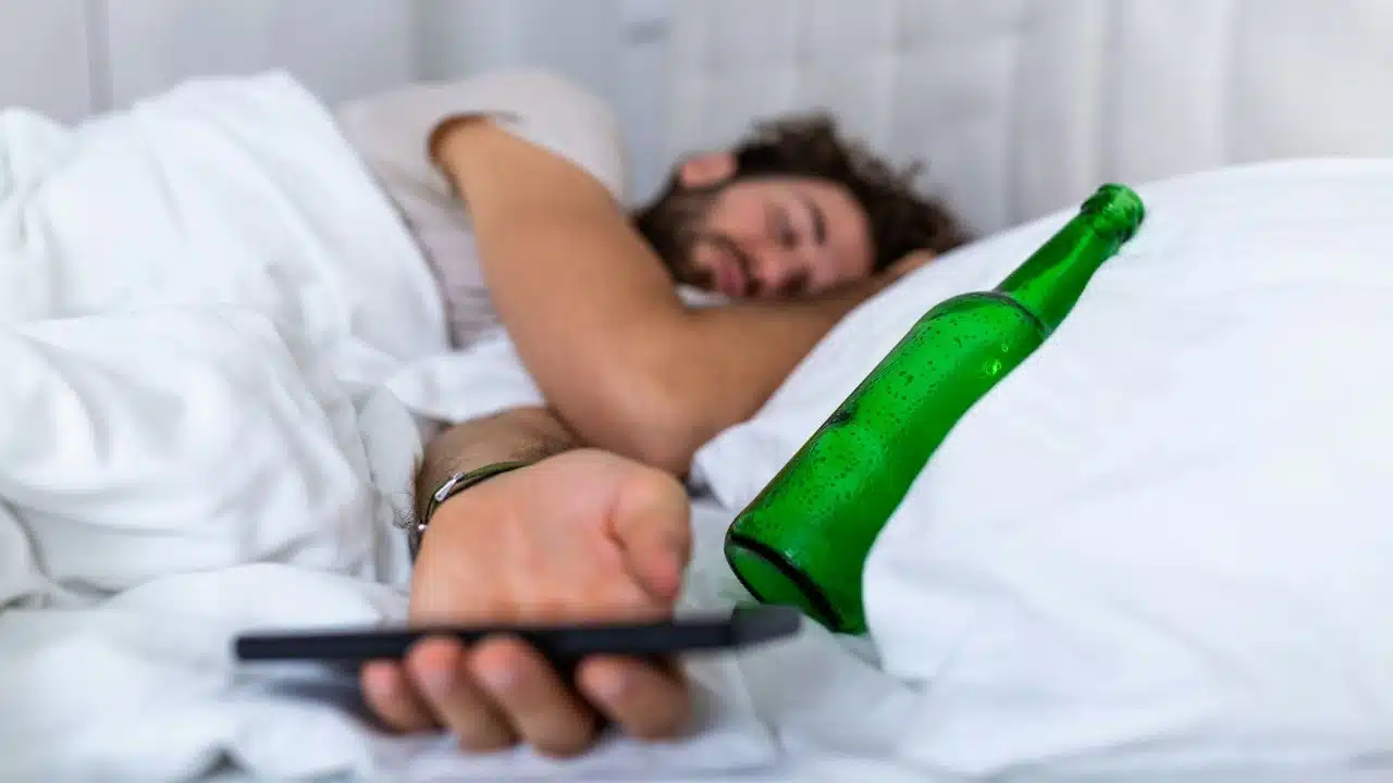 A man sleeps in bed next to a green glass bottle and a smartphone.